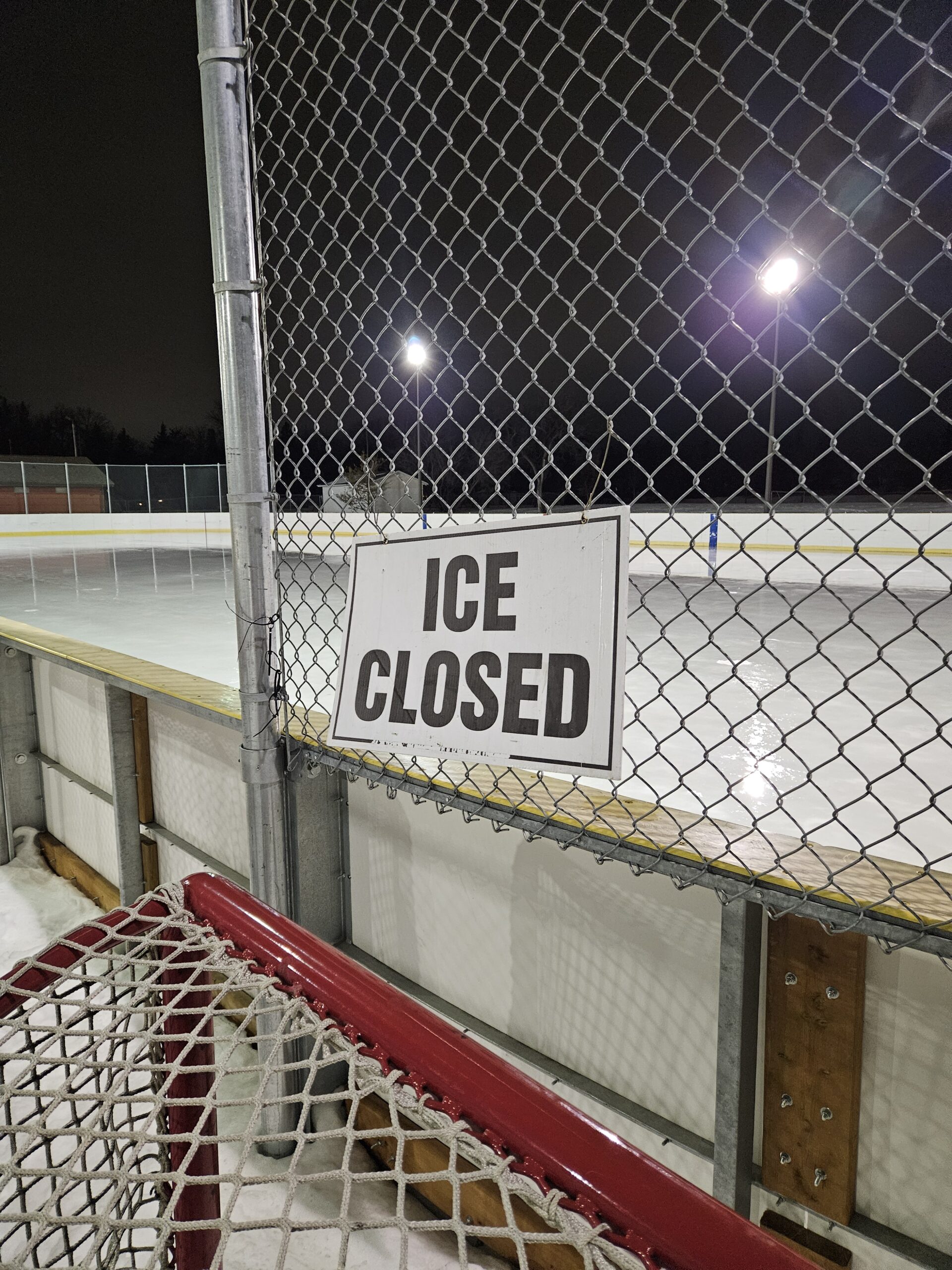 The Bourkevale south hockey rink at night. The rink is enclosed by a chain-link fence, and part of a hockey goal net is visible in the foreground, leaning against the fence. There is a sign attached to the fence that reads "ICE CLOSED", indicating that the rink is not currently open for skating. Bright lights illuminate the scene, likely from light poles positioned around the rink. The ice surface appears smooth and unoccupied, and there is some snow piled against the fence.