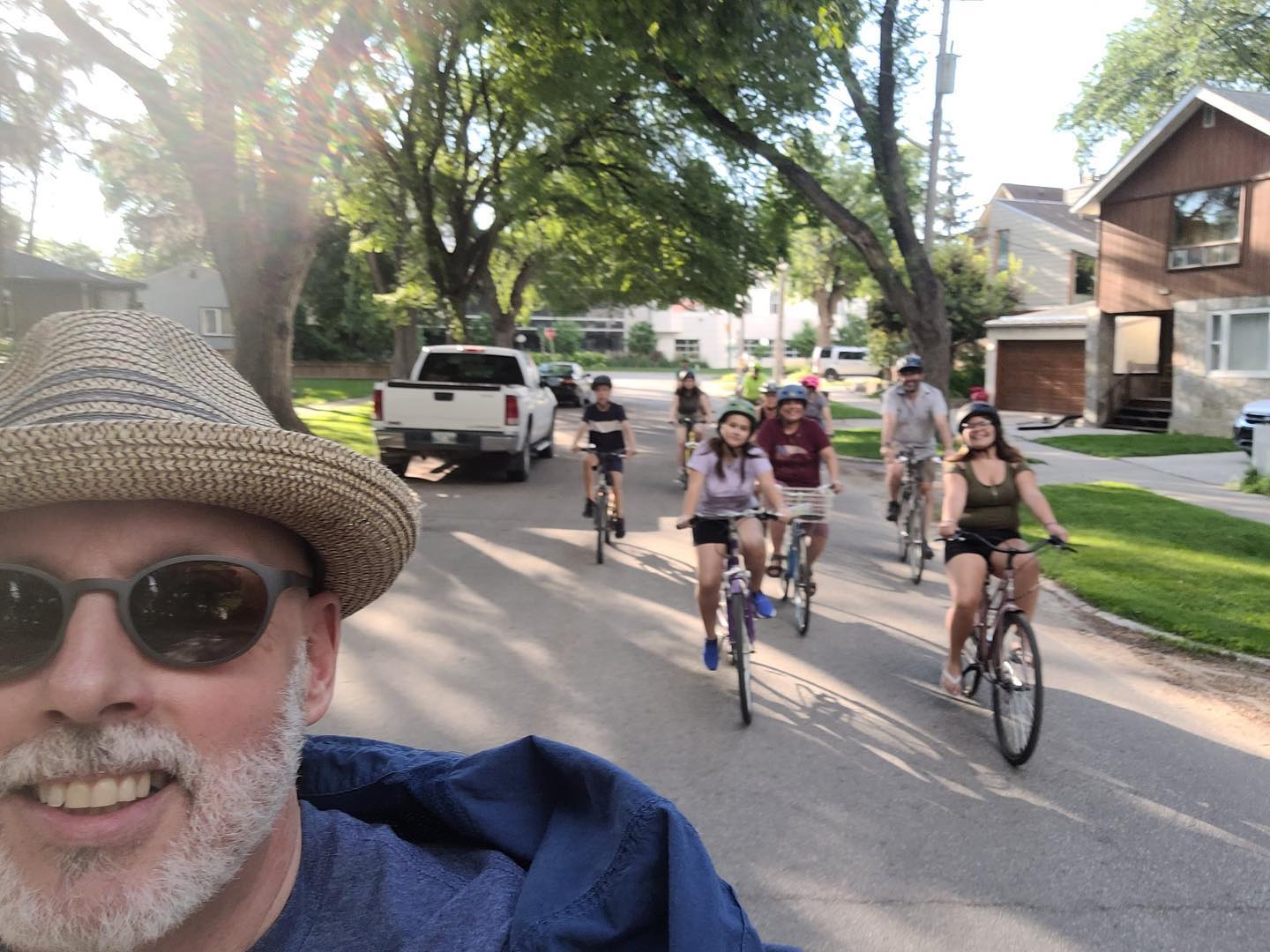 A group of people ride bicycles down a residential street. The person taking the selfie is in the foreground, wearing a straw hat and sunglasses, smiling at the camera. Behind them, several cyclists of various ages are riding in a line, enjoying the ride. The street is lined with trees, and there are houses and parked vehicles visible along the sides. The atmosphere appears to be casual and friendly, with everyone seemingly enjoying a group bike ride on a sunny day.
