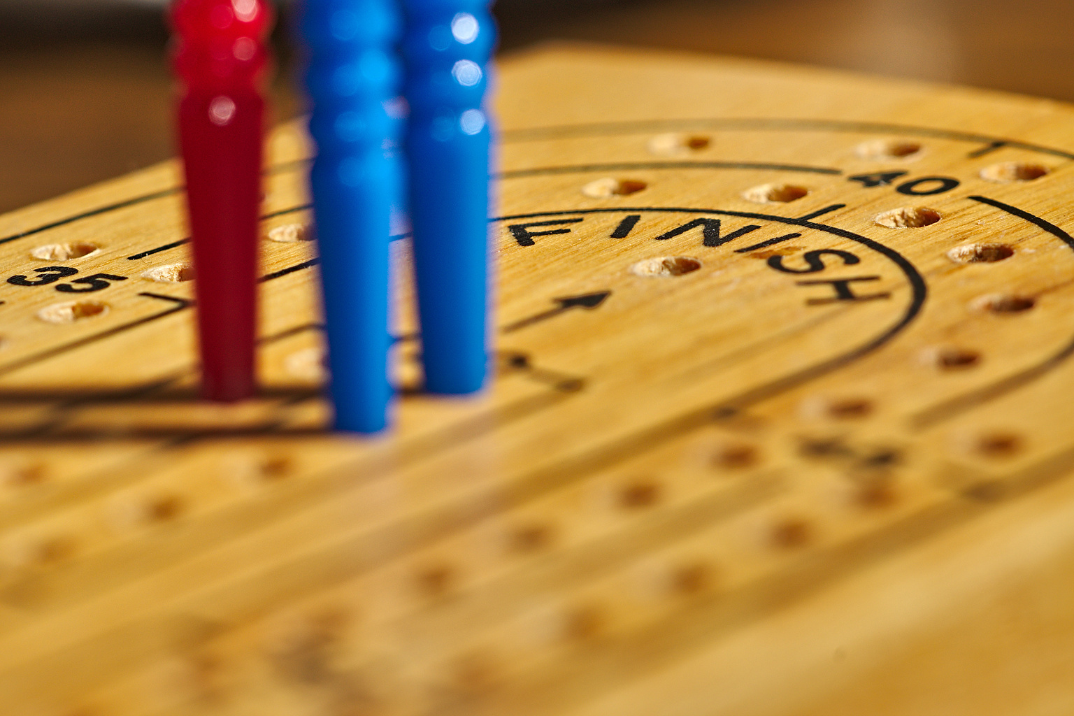 A close-up of a cribbage board with pegs. The board is made of wood and features a circular playing area with holes for pegs. Three pegs, two blue and one red, are positioned near the center of the board, close to the "FINISH" label.